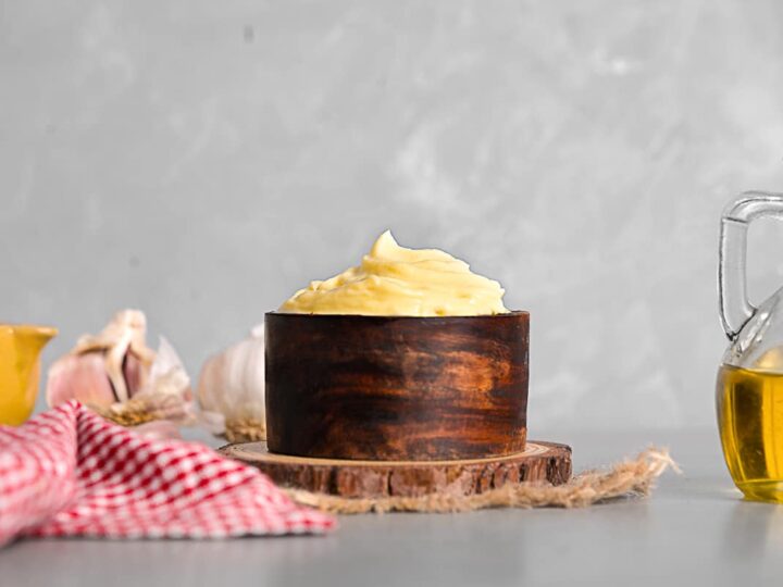 Creamy Spanish alioli garlic sauce in a wooden bowl, surrounded by garlic cloves, olive oil, and a checkered cloth, highlighting traditional Spanish cuisine.